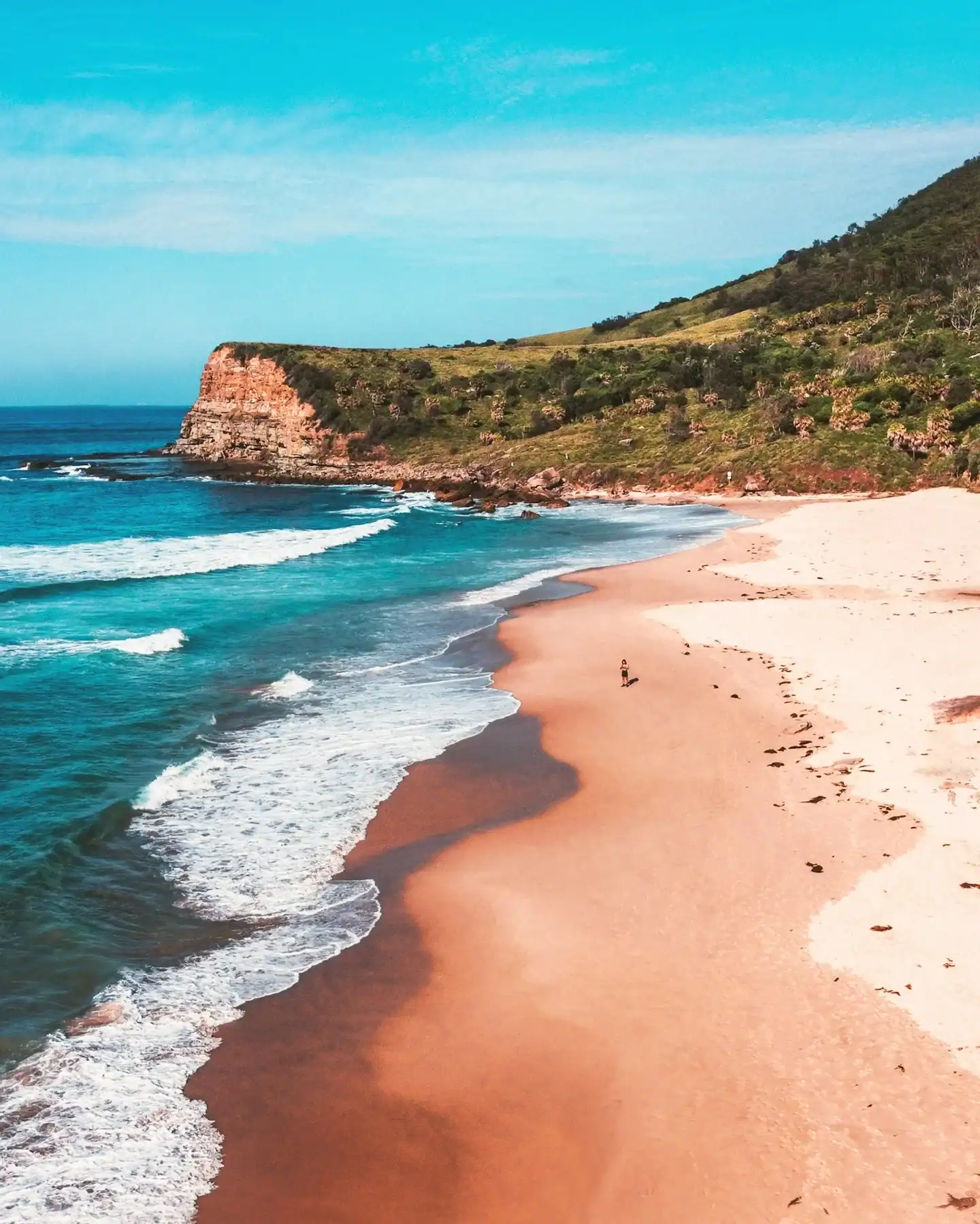 A person walking along the sand beneath a grassy coastal headland