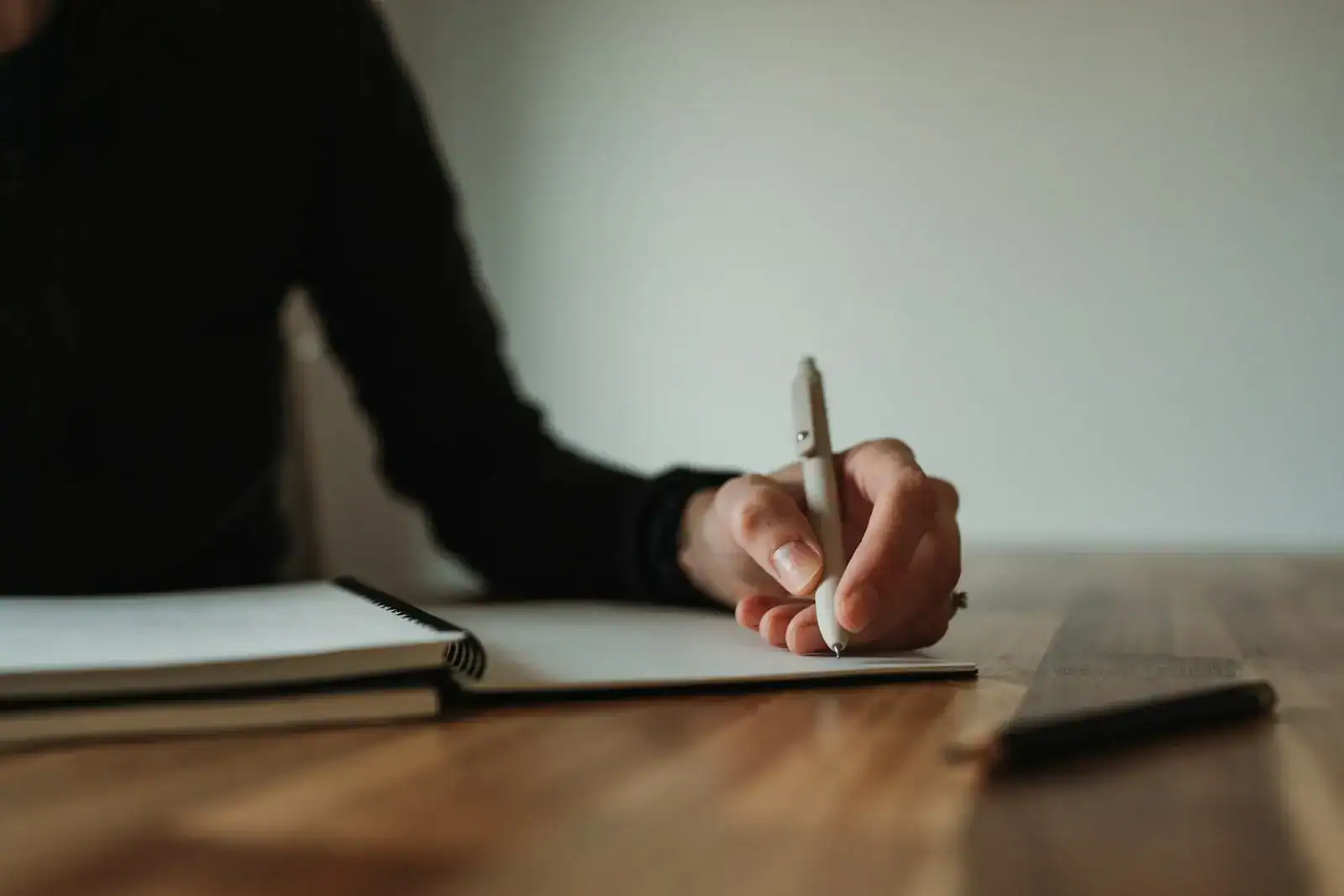 A hand taking notes in an open book on a wooden desk
